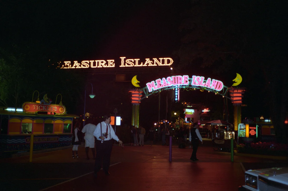 Illuminated Pleasure Island entrance arch beside a ticket booth at night, with visitors and staff nearby.